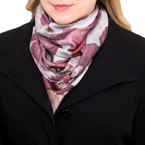 Woman wearing a silk scarf with overlapping floral images in red, berry, maroon, and light tones, and hand-hemmed edges, while wearing a black coat. 