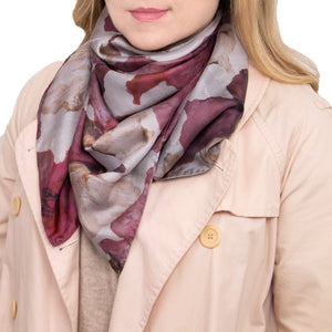 Woman wearing a silk scarf with overlapping floral images in red, berry, maroon, and light tones, and hand-hemmed edges, while wearing a light coat and a beige sweater.