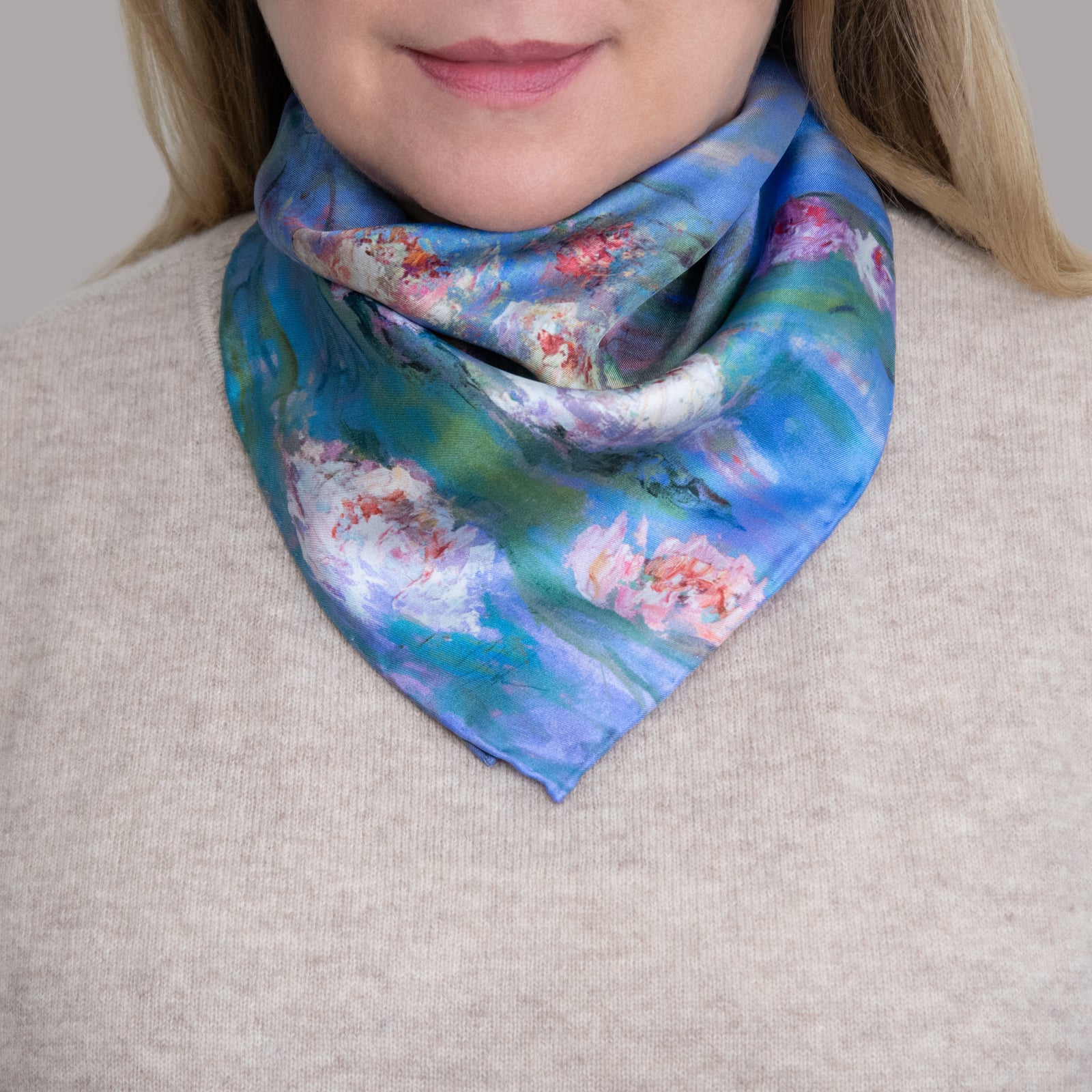 Woman in beige sweater wearing a small Floating Nympheas silk scarf around her neck, with pink, yellow, and white blossoms showing on blue water.