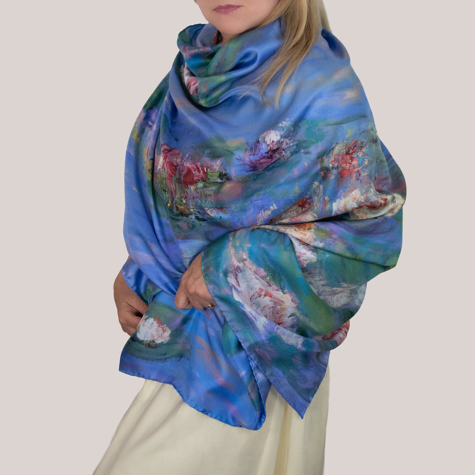 Woman in a light-colored dress wearing a large Floating Nympheas silk scarf as a wrap, showing white, yellow, and pink water lily blossoms and lily pads on a field of blue water. 