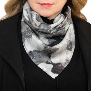 Woman wearing a medium-sized silk scarf in a floral pattern with black, white, and gray overlapping images and hand hemmed edges, along with a black top. 