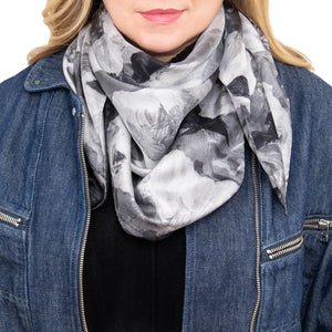 Woman wearing a silk scarf in a floral pattern with black, white, and gray overlapping images and hand hemmed edges, along with a jean jacket and black top. 
