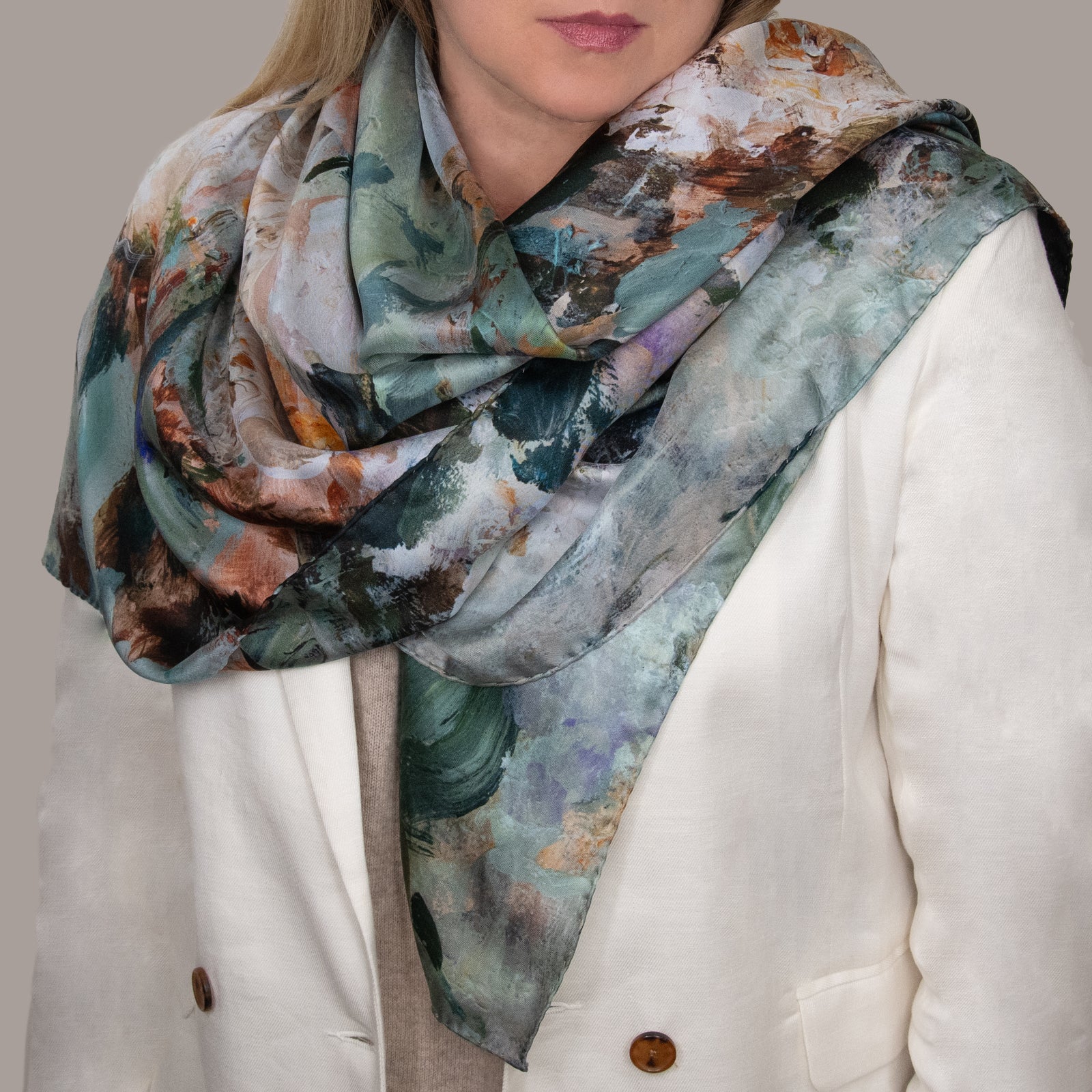 Woman in a white coat and beige sweater wearing a large scarf with impressionist designs in green, white, brown, and purple. 
