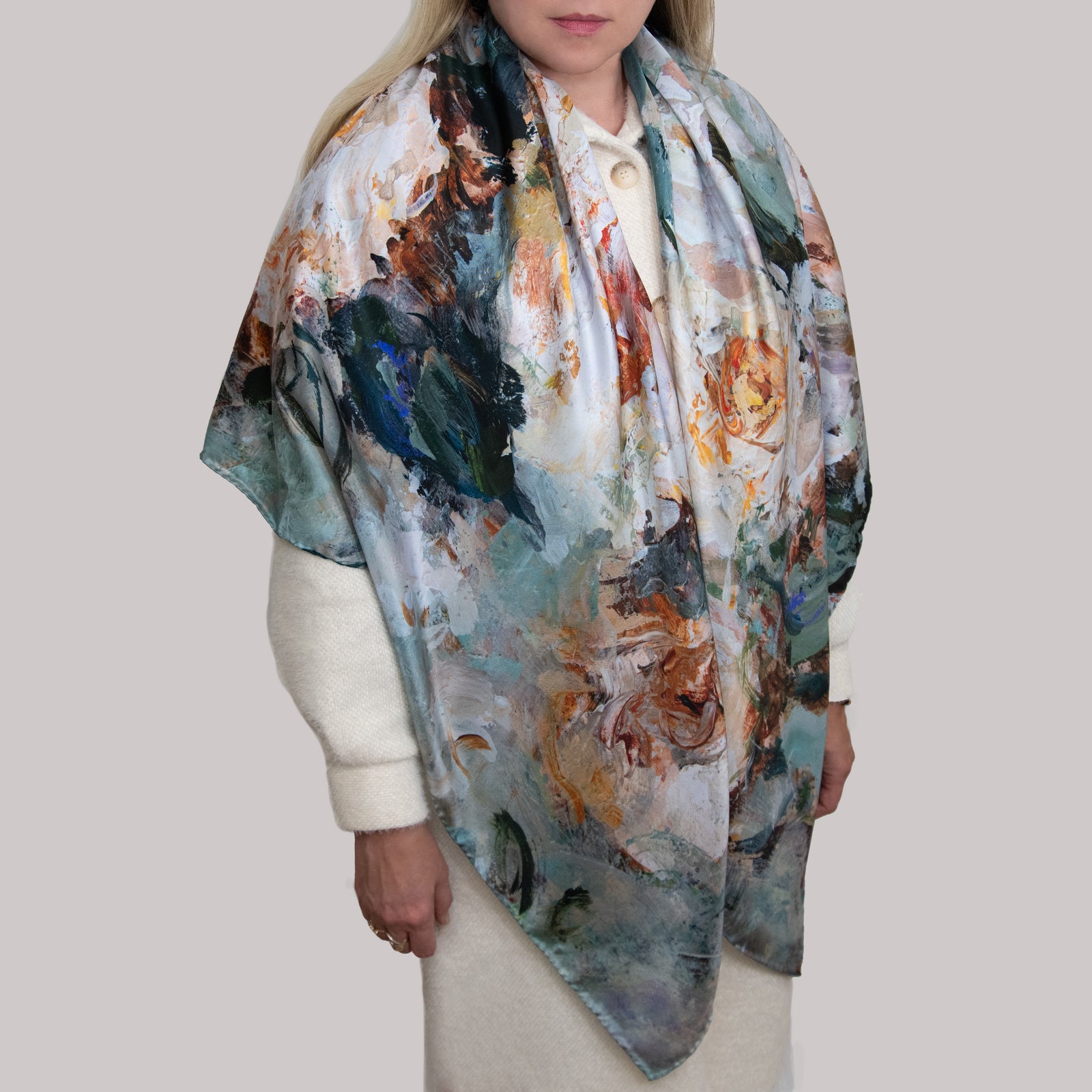 Woman wearing a large impressionist scarf or wrap with white and yellow roses on a green and dark background, draped across shoulders of her white coat and hanging down below her waist. 