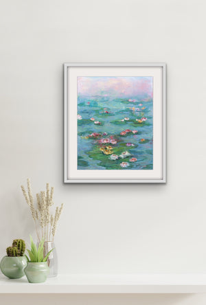 Sunrise over the Pond, archival fine art print by Oksana Johnson, shown framed on a wall with a shelf and plants below — soft morning light dissolving over a lily-covered pond in blush pink, teal, and warm coral