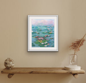 Sunrise over the Pond, archival fine art print by Oksana Johnson, shown on a beige wall above a wooden shelf with decorative items — soft morning light dissolving over a lily-covered pond in blush pink, teal, and warm coral