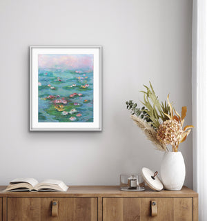 Sunrise over the Pond, archival fine art print by Oksana Johnson, shown in a gray frame on the wall above a wooden cabinet with a book and vase — soft morning light dissolving over a lily-covered pond in blush pink, teal, and warm coral