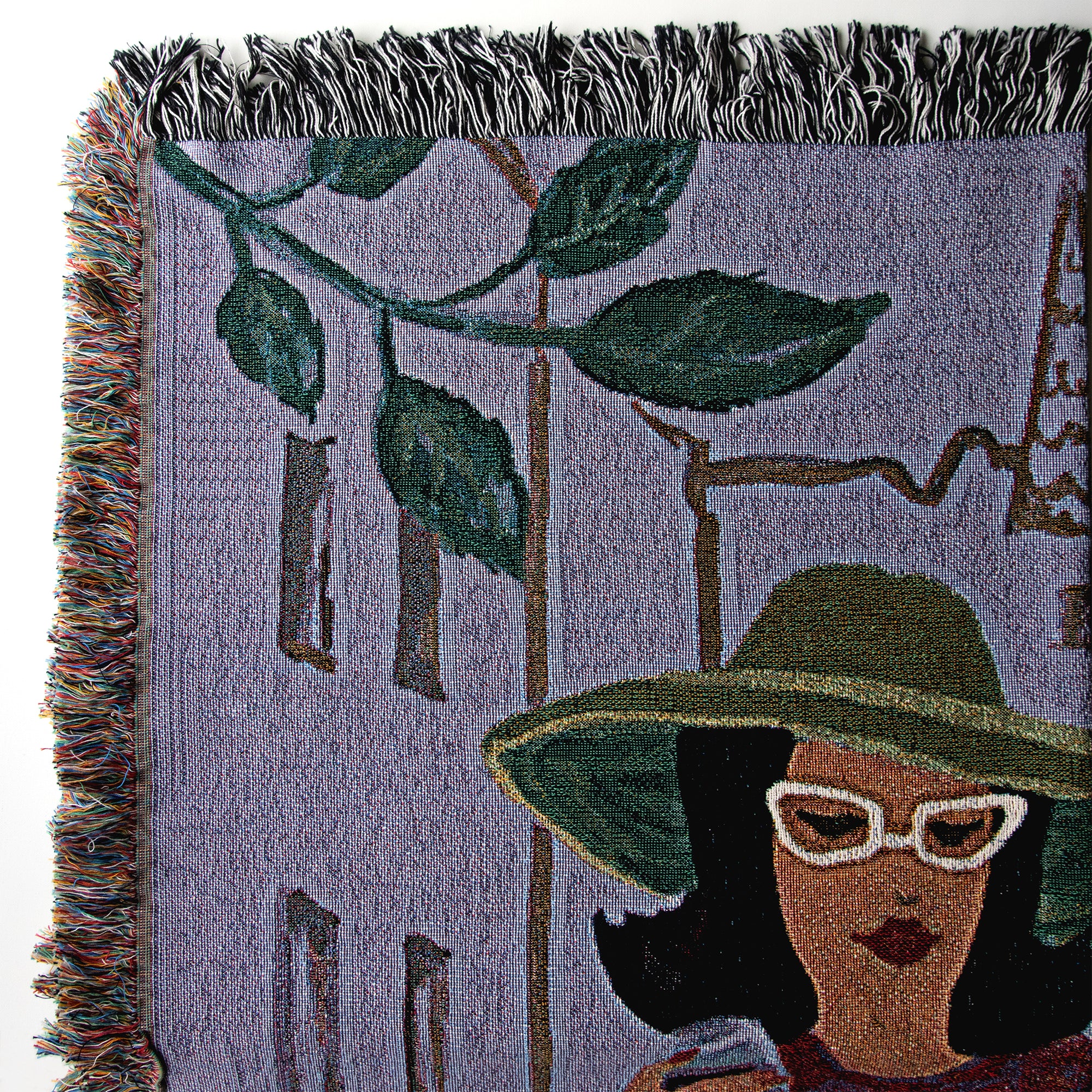 Details from upper left 
corner of a woven blanket with a design of a woman wearing a green hat and white sunglasses, with plants and buildings in the background, showing close-up of the colored fringes on one side and the black-and-white fringes on the top. 