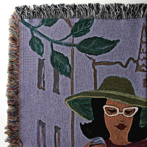 Details from upper left 
corner of a woven blanket with a design of a woman wearing a green hat and white sunglasses, with plants and buildings in the background, showing close-up of the colored fringes on one side and the black-and-white fringes on the top. 