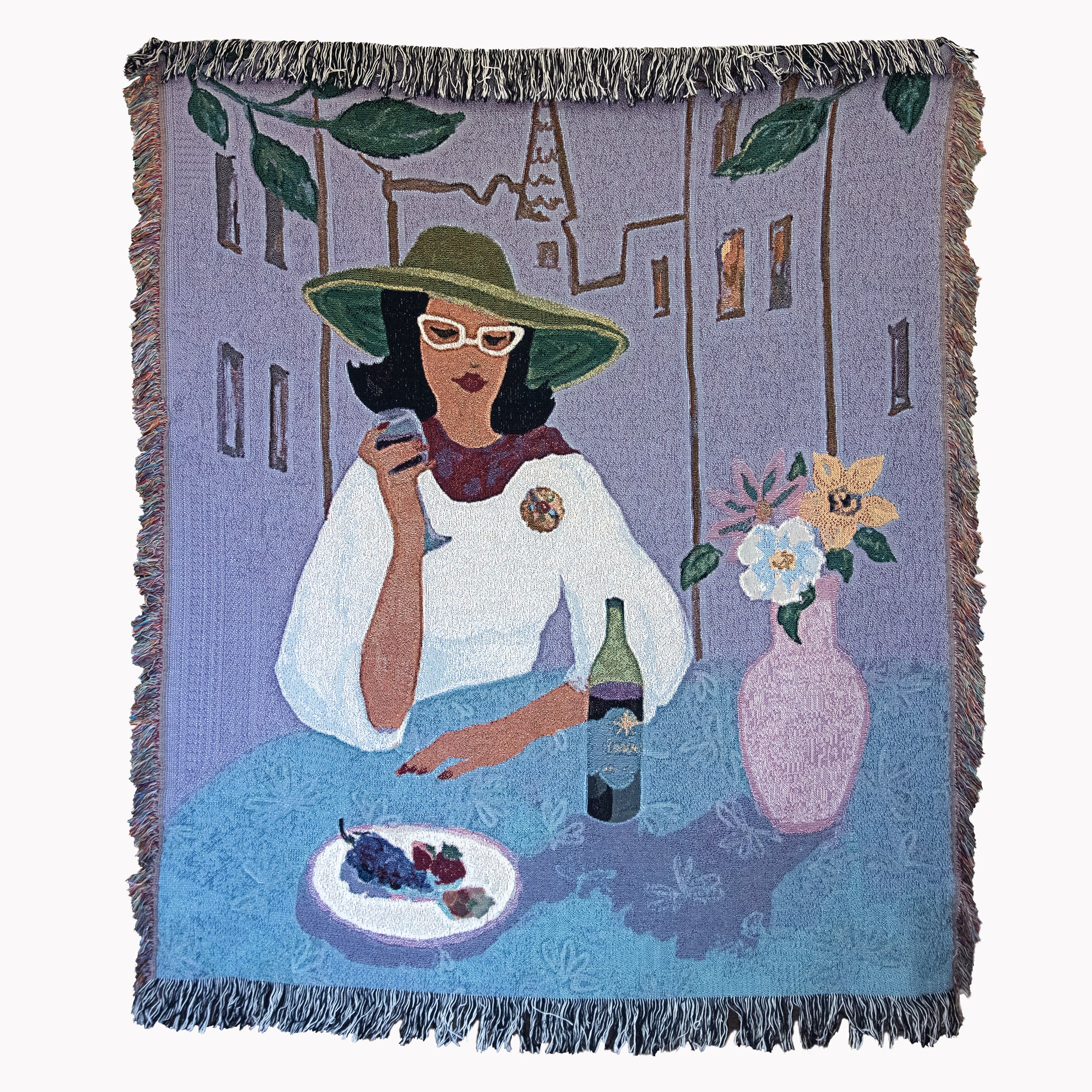 Woven blanket showing a stylish woman in a white top, green hat, and glasses sitting at a table scene with wine, fruit, and flowers, against a backdrop of buildings, with fringed edges.