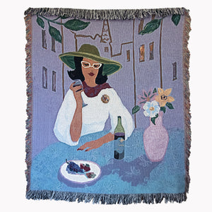 Woven blanket showing a stylish woman in a white top, green hat, and glasses sitting at a table scene with wine, fruit, and flowers, against a backdrop of buildings, with fringed edges.
