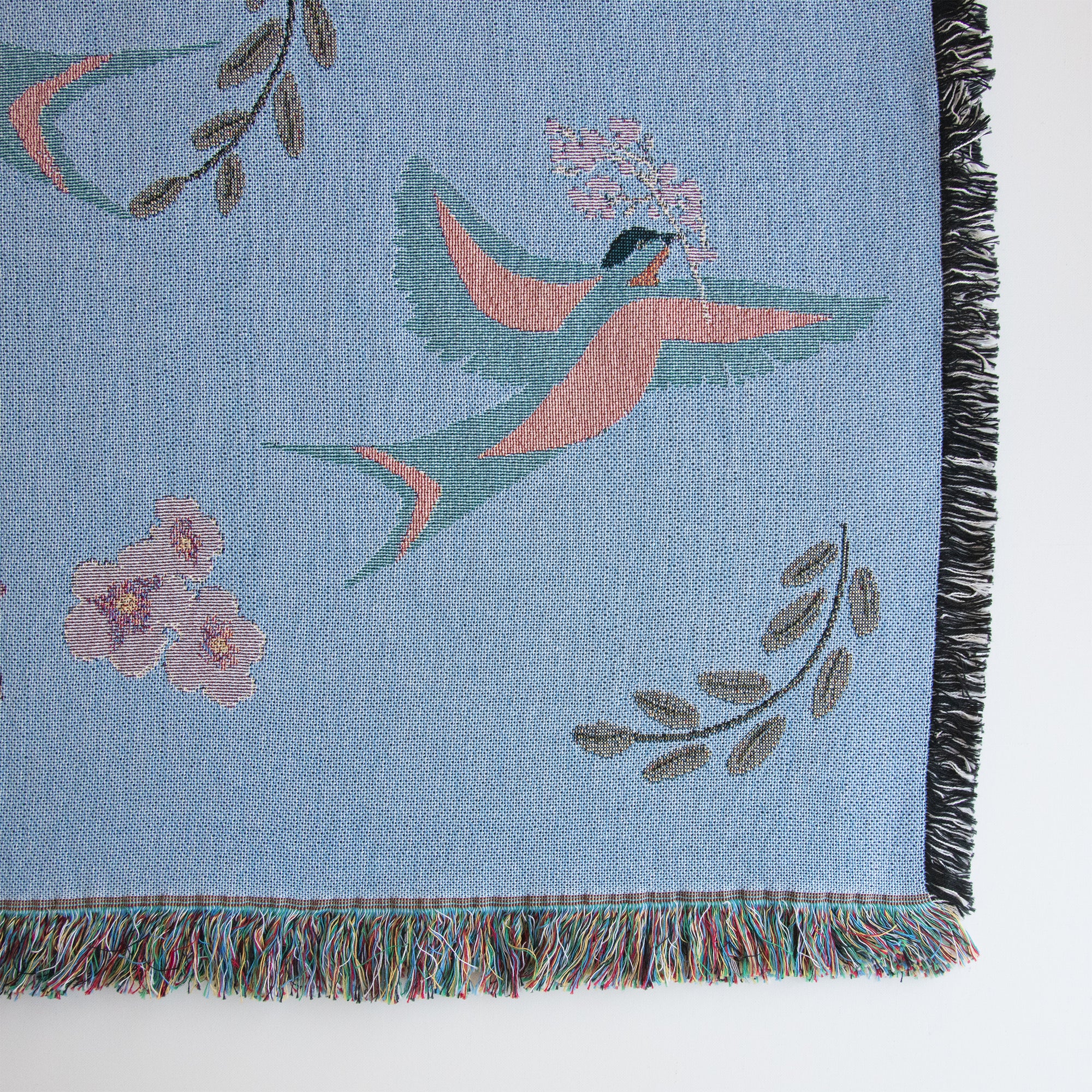 Close-up of lower right corner of decorative blanket with blue birds and flower pattern, showing fringed edges and details of the weave. 