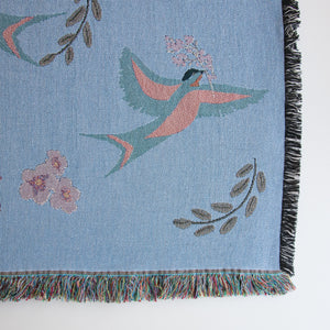Close-up of lower right corner of decorative blanket with blue birds and flower pattern, showing fringed edges and details of the weave. 