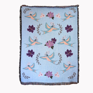 Decorative fine art blanket with blue birds and flower pattern woven into a light blue background, with fringed edges.