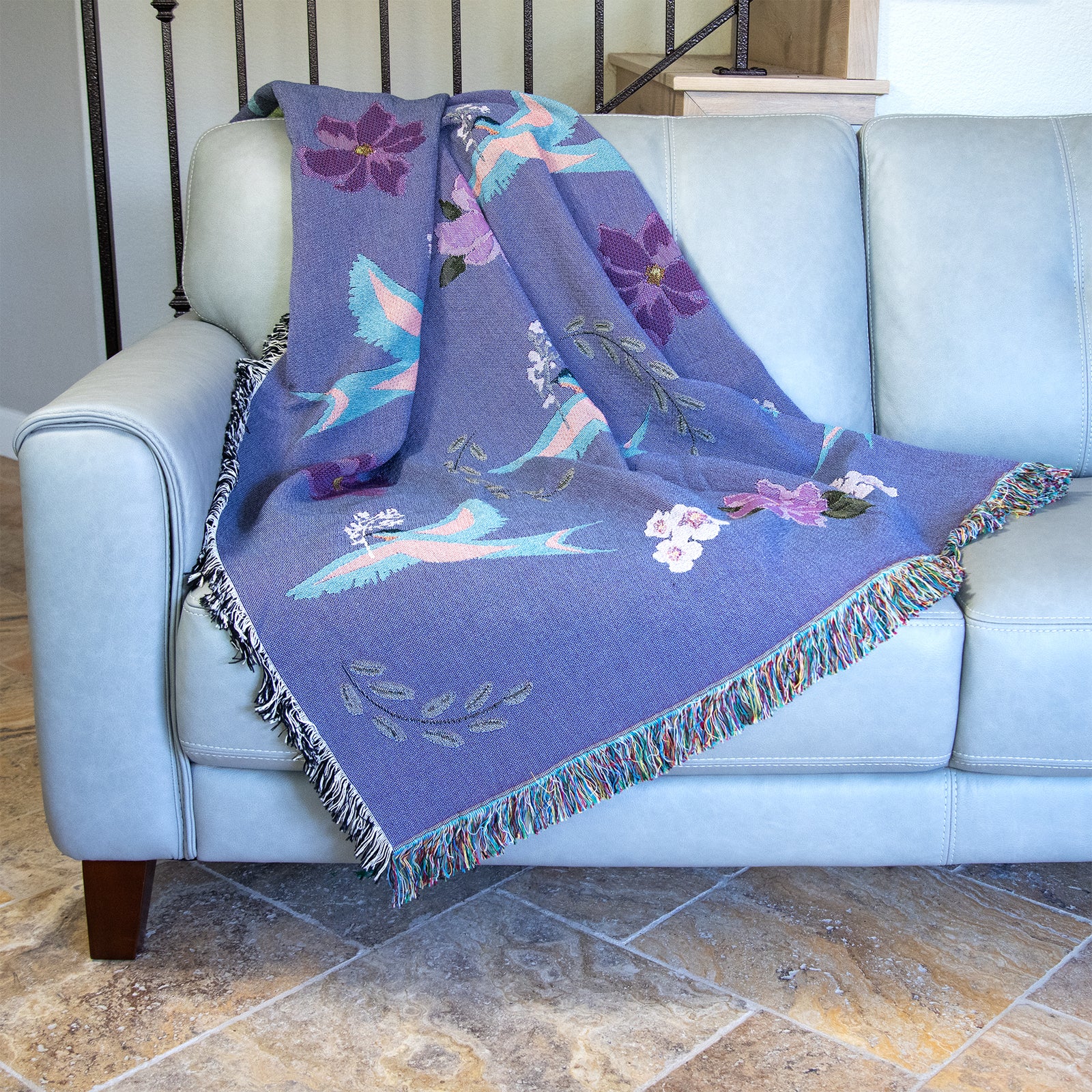 Decorative blanket with light blue birds and flower pattern woven into a purple background, with fringed edges.