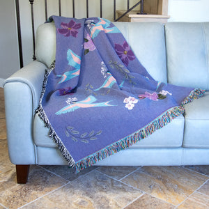 Decorative blanket with light blue birds and flower pattern woven into a purple background, draped on a light blue-grey sofa