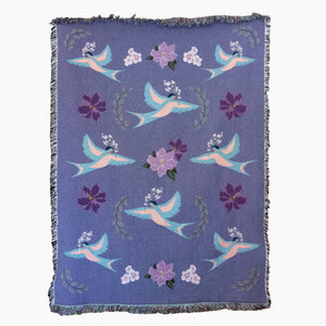 Decorative blanket with light blue birds and flower pattern woven into a purple background, with fringed edges.